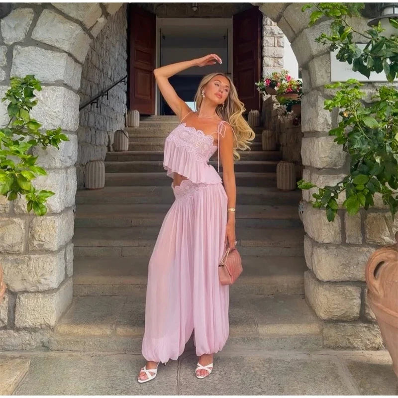 A woman posing in a light pink two-piece outfit consisting of a ruffled crop top and matching wide-leg pants, standing on stone steps between arches.